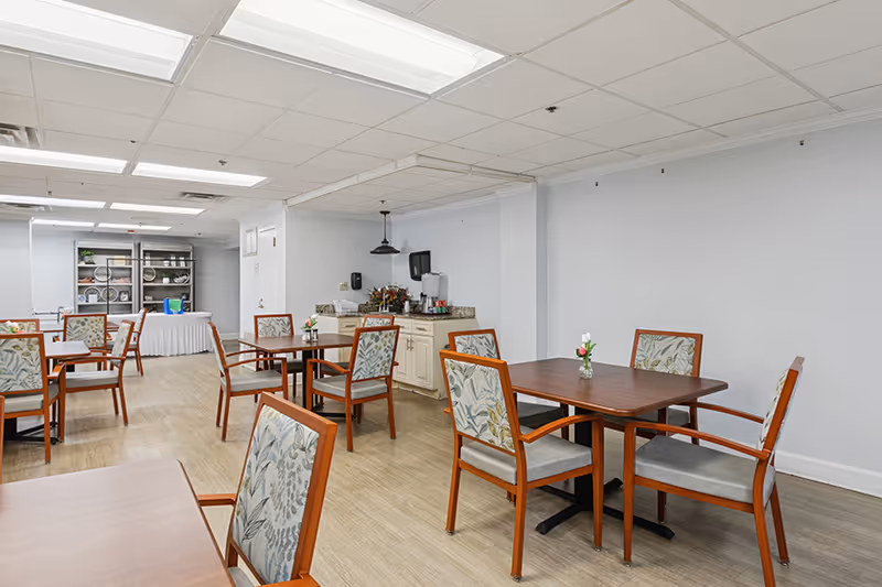 A dining room with several wooden tables and chairs with floral-patterned cushions. The room has light-colored walls and a light wood floor. In the background, there is a counter with a coffee station and a shelving unit with plates and other dining items.