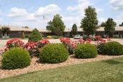 View of a landscaped outdoor area with neatly trimmed bushes and blooming red flowers in the foreground, a paved driveway, and a single-story building with an American flag on a flagpole in the background under a partly cloudy sky.