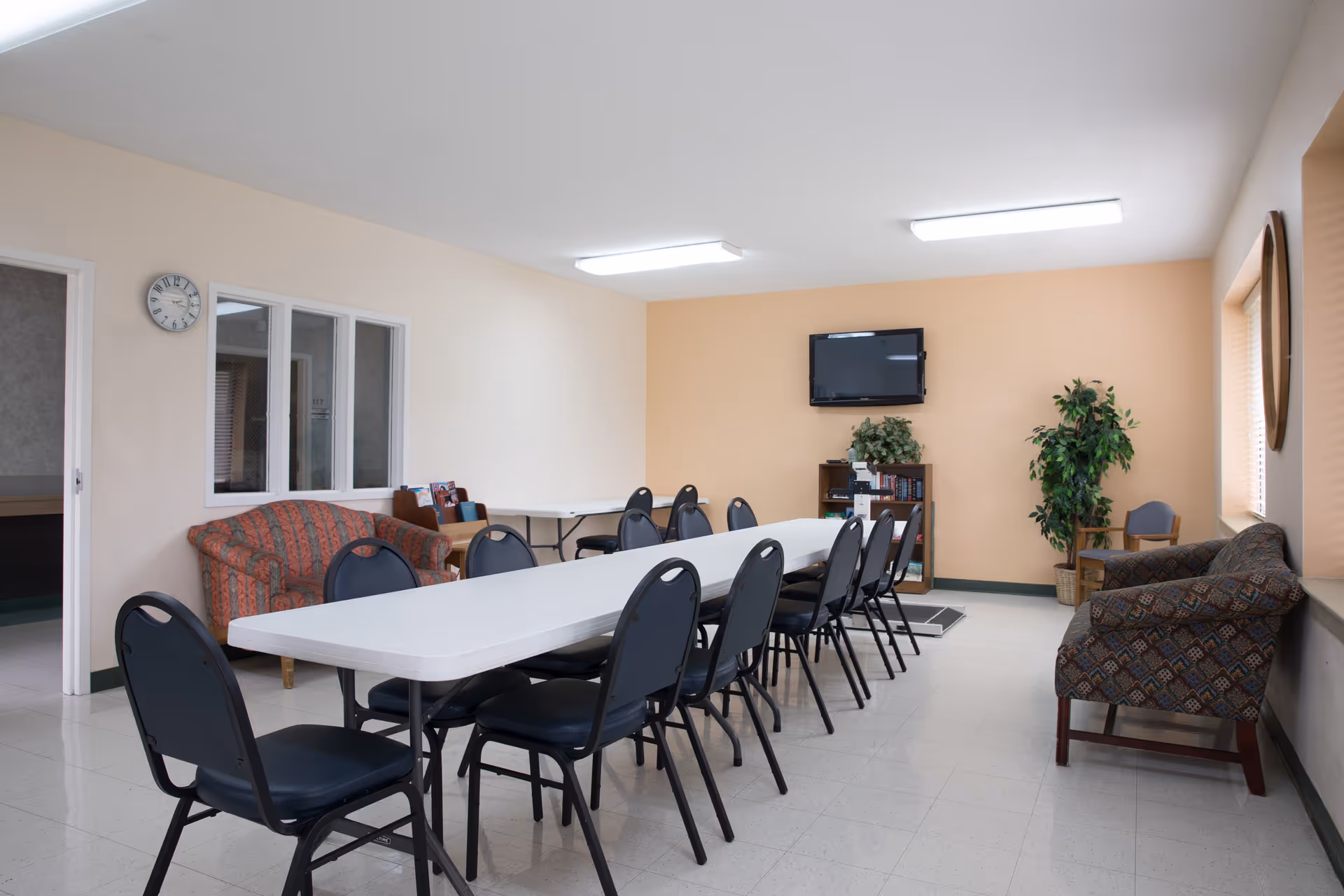 A community room with a long white table surrounded by black chairs. The room has beige walls, a wall-mounted TV, two potted plants, a bookshelf with books, and two patterned sofas. There is a clock on the wall and a window looking into another room.