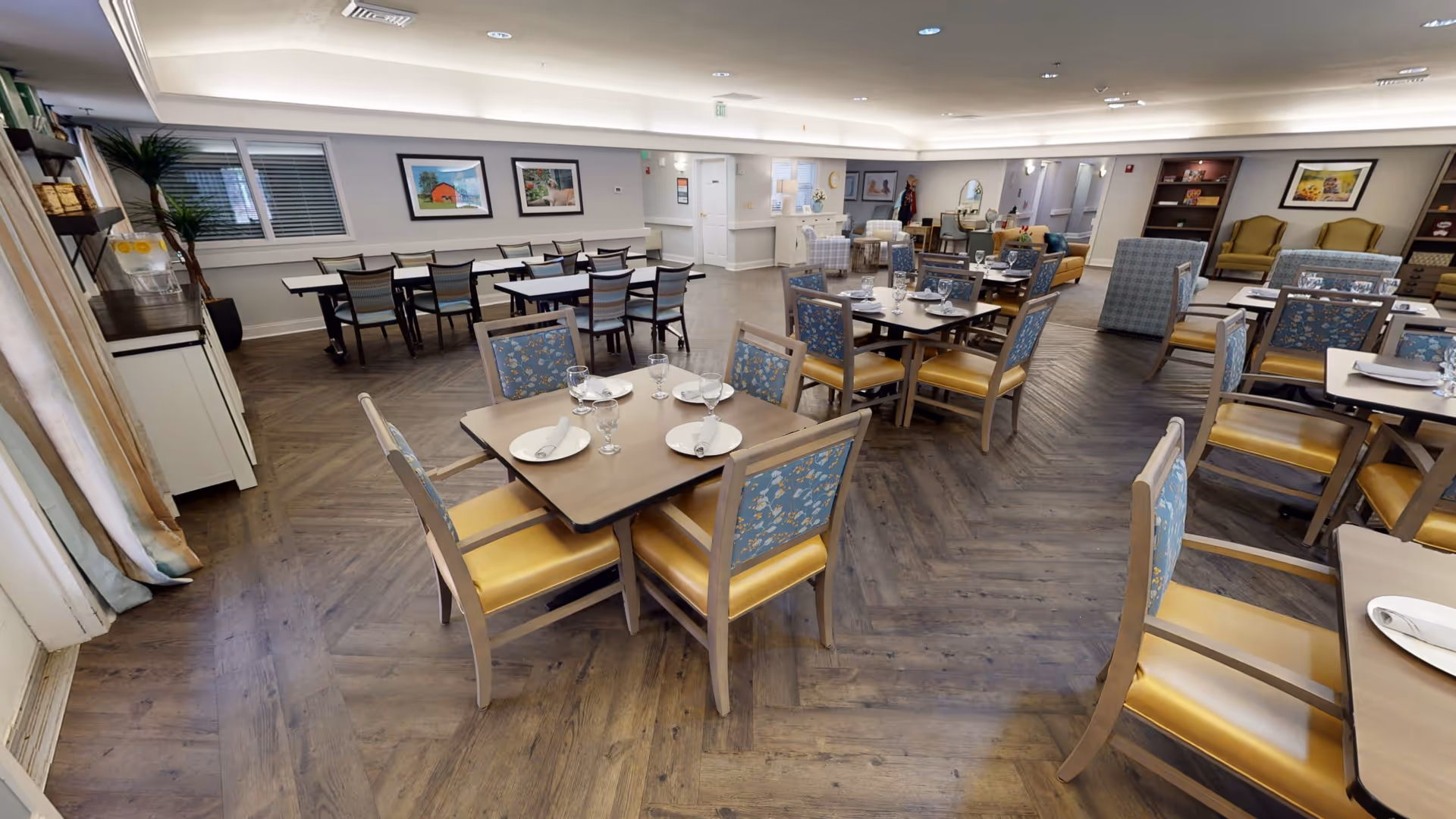 A spacious dining room with multiple tables and chairs arranged neatly. Each table is set with plates, napkins, and glasses. The room has wooden flooring, soft lighting, and framed artwork on the walls. There are also some lounge chairs and bookshelves in the background.