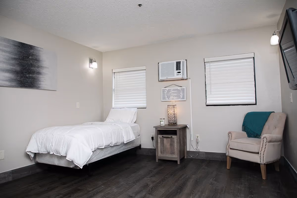 Simple furnished bedroom with a single bed, nightstand and lamp, armchair, and a wall air conditioner.