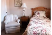 A bedroom with floral bedding, wooden headboard, nightstand, lamp, and a white armchair.