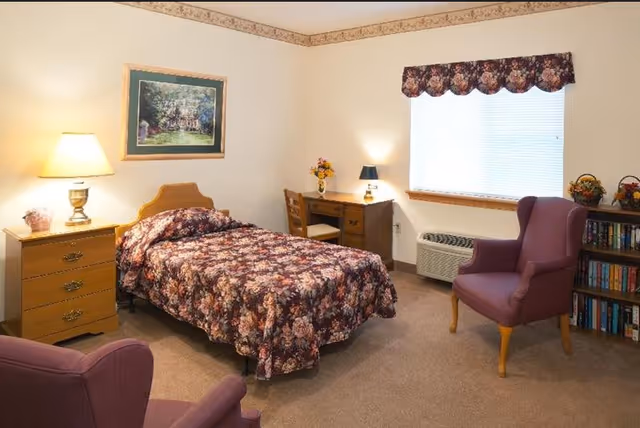 A senior living bedroom with a single bed covered in a floral bedspread, a wooden nightstand with a lamp, a wooden desk with a chair and a small lamp, two purple armchairs, a bookshelf filled with books, a window with blinds and a floral valance, and a framed picture on the wall.