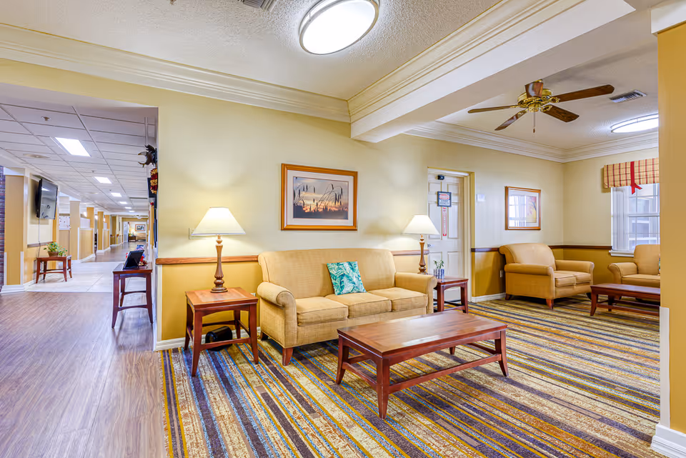 Well-lit senior living common area with sofas, armchairs, coffee tables, lamps, framed artwork, and a striped carpet.