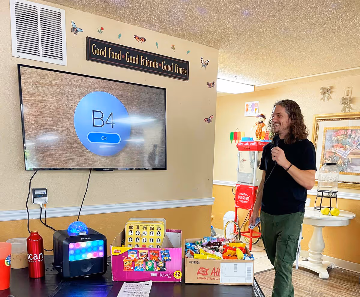 A man holding a microphone stands beside a wall-mounted TV and a table of snacks, bingo supplies, and a popcorn machine in a decorated activity room.