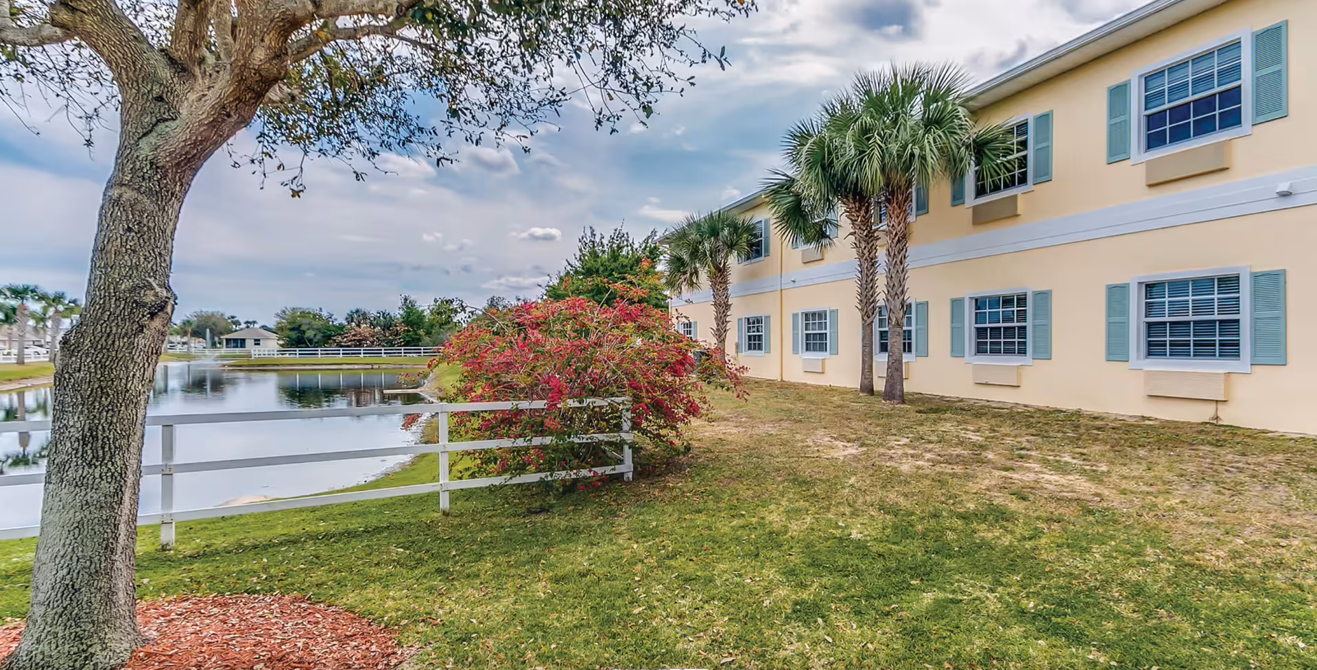 View of the exterior side of a two-story yellow building with blue window shutters, palm trees, and a grassy area leading to a white fence bordering a pond. The sky is partly cloudy.