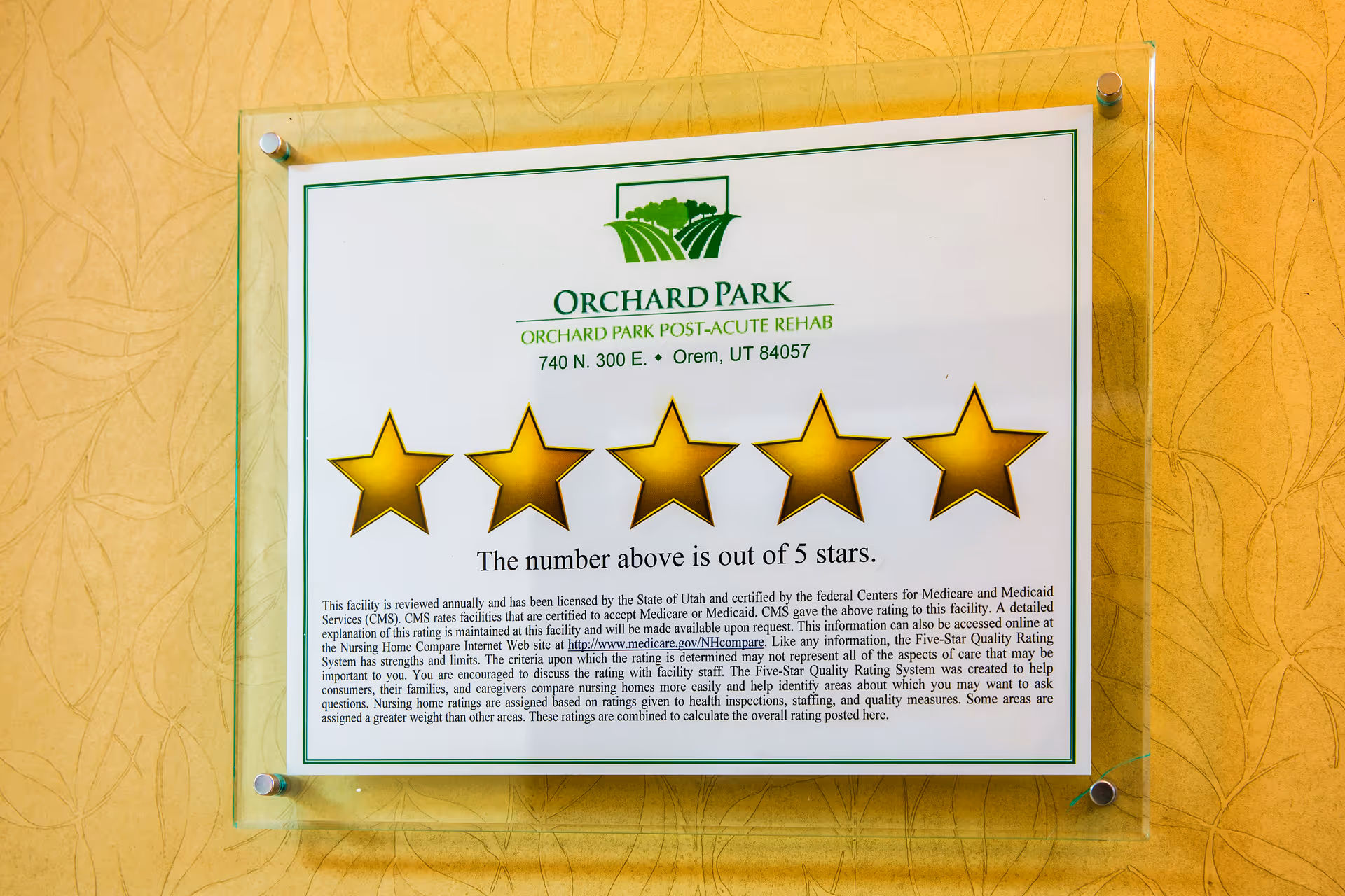 A framed sign on a yellow textured wall displaying a five-star rating for Orchard Park Post-Acute Rehab located at 740 N. 300 E., Orem, UT 84057. The sign explains that the facility is rated 5 out of 5 stars by the Centers for Medicare and Medicaid Services (CMS) and provides information about the rating system and where to find more details online.