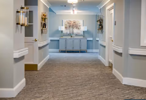 Carpeted interior hallway with light gray walls, wall sconces and doors, and a decorative console table with artwork at the far end.