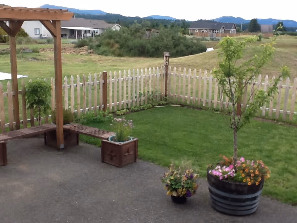 Patio and small fenced backyard with a wooden pergola bench, potted flowers, a grassy lawn, and houses and hills in the distance.