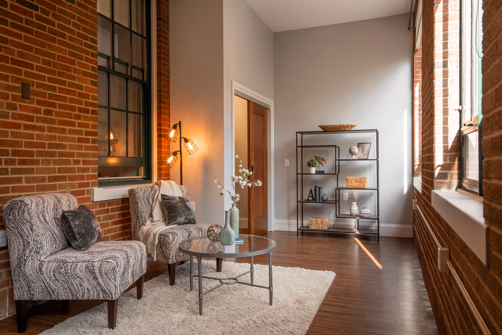 Sunlit lounge with two patterned armchairs around a glass coffee table, a metal shelving unit, and exposed brick walls with large windows.