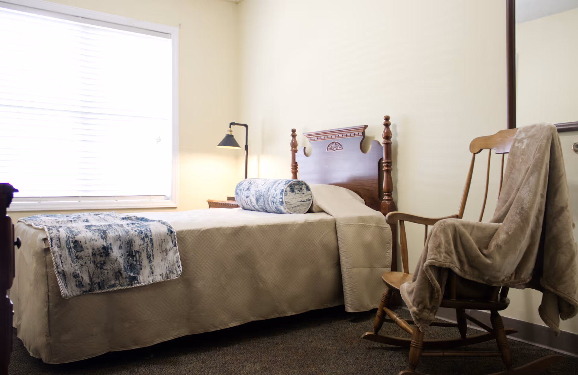A neatly made single bed with a beige bedspread and a blue patterned bolster pillow and throw blanket, positioned next to a window with blinds. A wooden rocking chair with a beige blanket draped over it is placed beside the bed. A small bedside table with a black lamp is also visible.