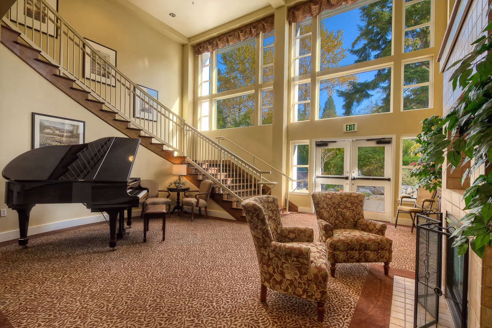 A bright and spacious living room area with large floor-to-ceiling windows letting in natural light. The room features a grand piano on the left, two patterned armchairs facing a fireplace on the right, and a staircase with a metal railing leading to an upper floor. There are framed pictures on the wall and a small table with chairs near the staircase. Outside the glass doors, greenery and trees are visible.