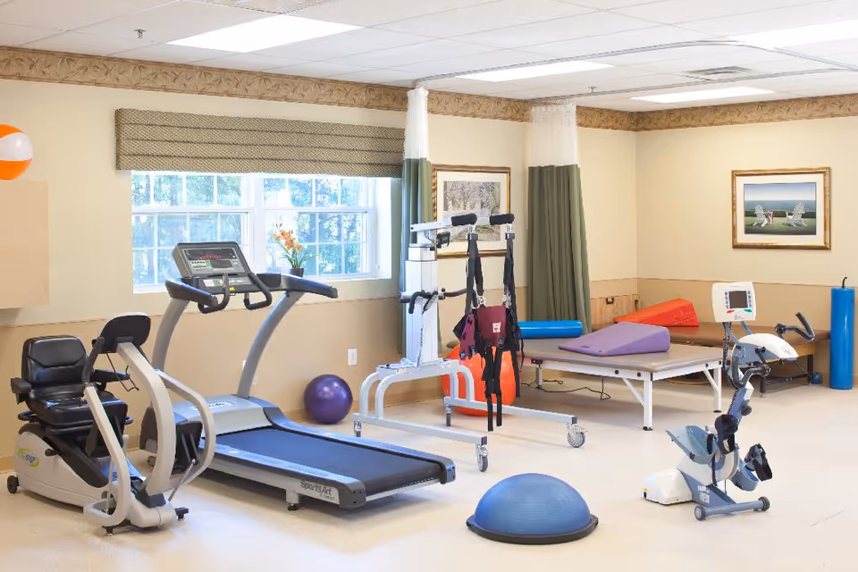 A bright and clean physical therapy or exercise room with various fitness equipment including a recumbent bike, treadmill, stationary bike, therapy table with cushions, exercise balls, and a gait training device. The room has large windows with beige blinds, framed artwork on the walls, and a beige and light brown color scheme.