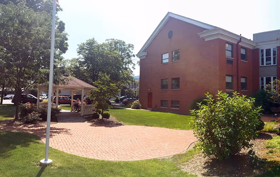 Outdoor view of Presbyterian Village at Hollidaysburg showing a brick building, a gazebo with people sitting inside, a flagpole, trees, bushes, and a paved walkway on a sunny day.