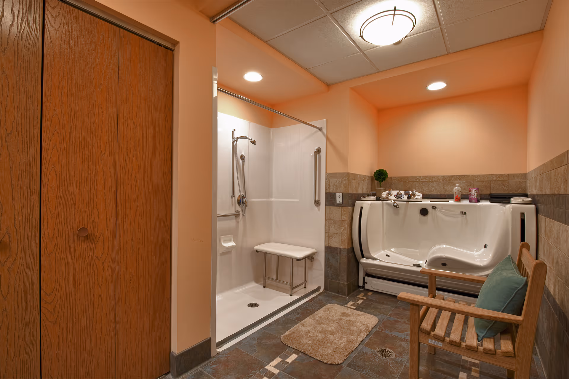 A bathroom with a walk-in shower featuring a foldable seat and grab bars, a large accessible bathtub with controls on the side, a wooden chair with a green cushion, and tiled floor and walls. The walls are painted peach and the ceiling has recessed lighting.