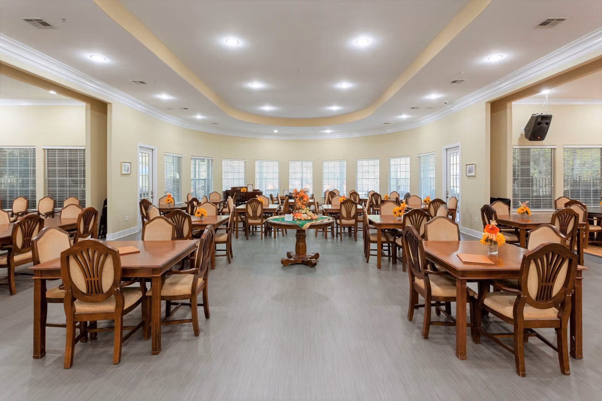 A spacious dining room with multiple wooden tables and chairs arranged neatly. Each table has a small floral centerpiece with orange flowers. The room has large windows along the back wall allowing natural light to fill the space. The ceiling features recessed lighting and a curved design. The floor is covered with light-colored wood or laminate flooring.