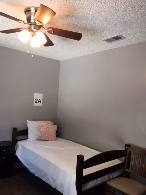 A small bedroom with a single bed made with white sheets, a white pillow, and a pink decorative pillow. There is a wooden chair next to the bed and a ceiling fan with lights above. The walls are painted gray, and a sign with '2A' is attached to the wall above the bed.