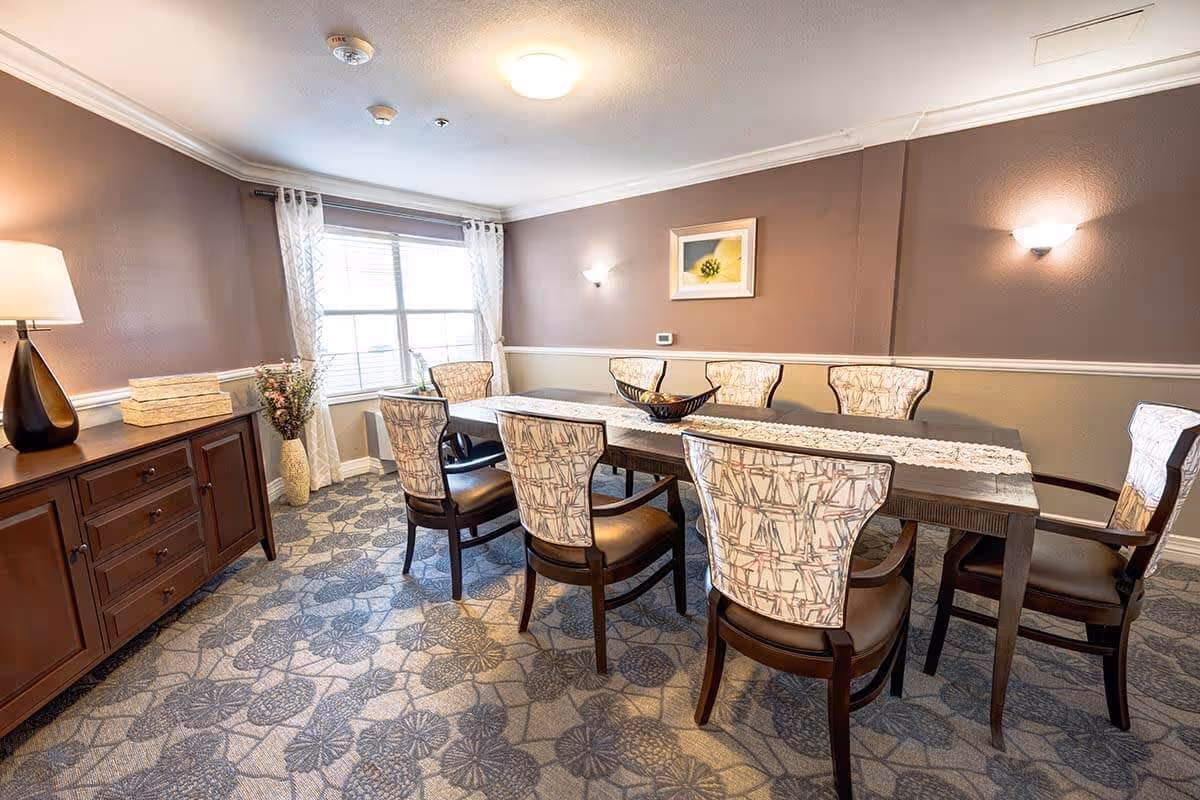 A dining room with a rectangular wooden table surrounded by eight upholstered chairs with patterned backs and dark wooden legs. The room has a patterned carpet, a sideboard with a lamp and decorative items, a large window with sheer curtains, and soft wall lighting. A framed picture hangs on the wall above the table.