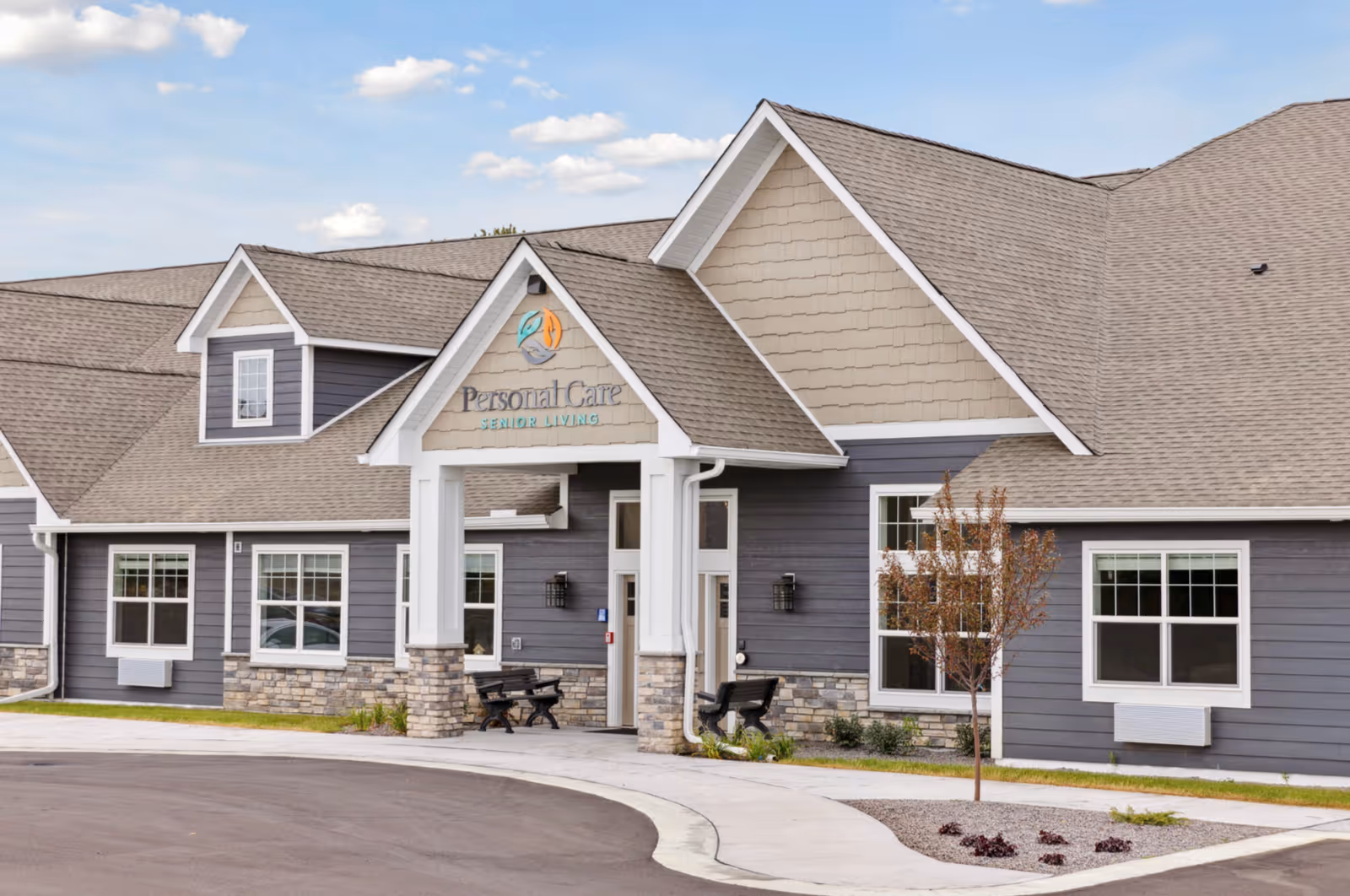Exterior view of Personal Care Senior Living - Andover facility showing a large building with gray and beige siding, stone accents, multiple windows, and a covered entrance with benches on either side. The sky is partly cloudy.