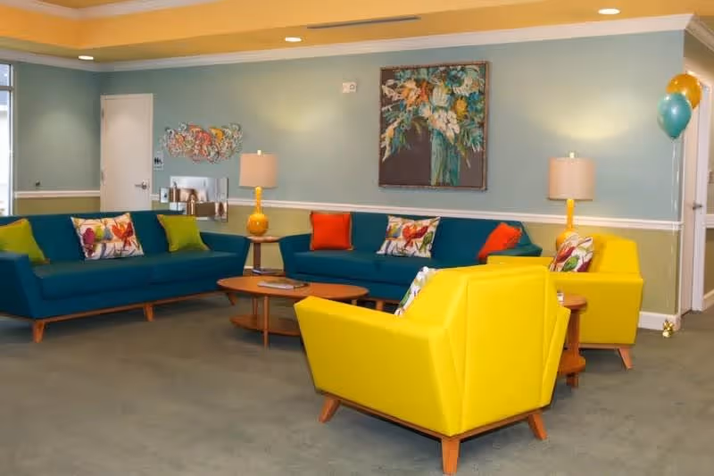 Bright communal lounge with teal sofas, yellow armchairs, wooden coffee tables and colorful pillows under wall art.