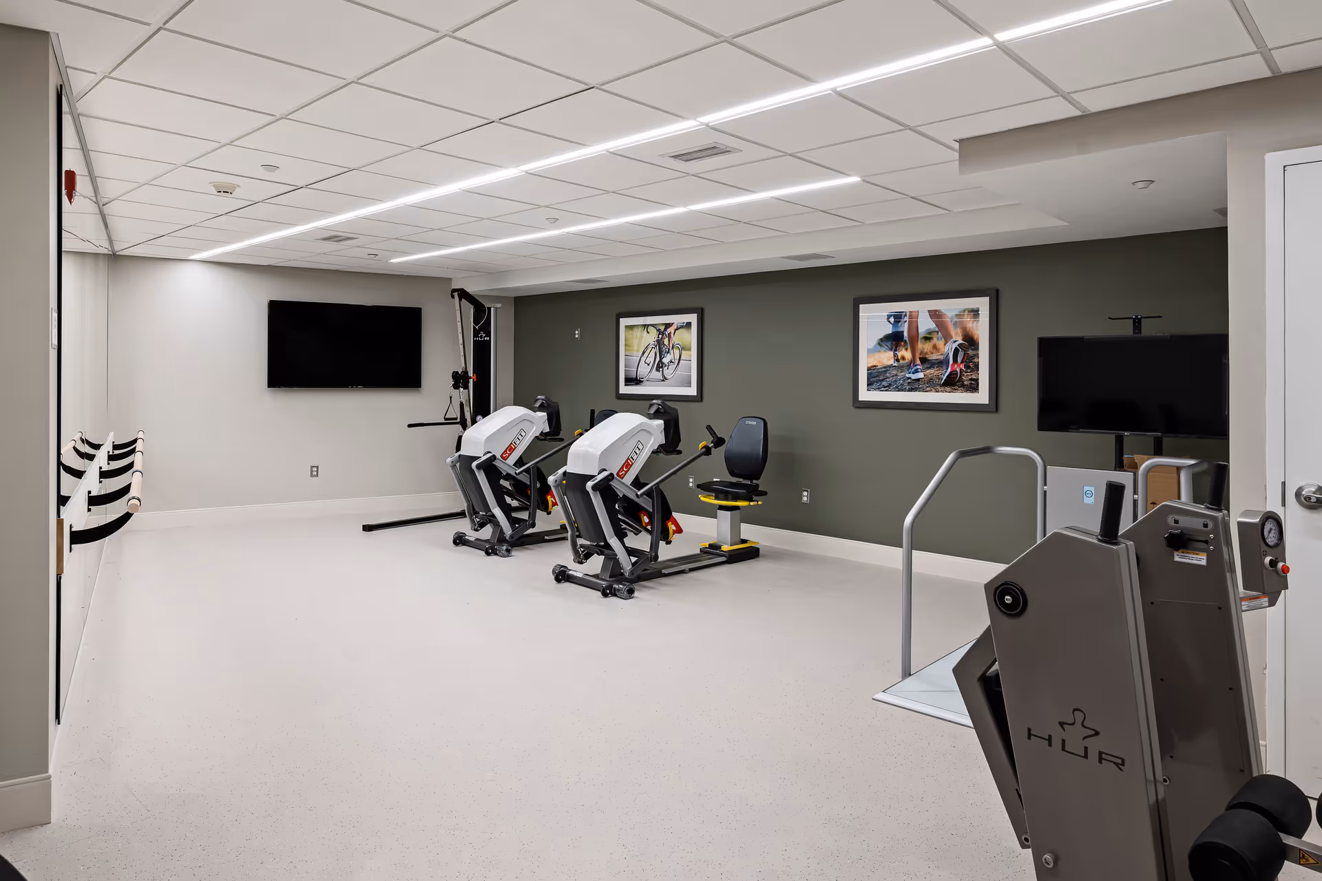 A bright senior facility exercise room with multiple recumbent bikes, wall-mounted TVs, framed photos, and handrails.