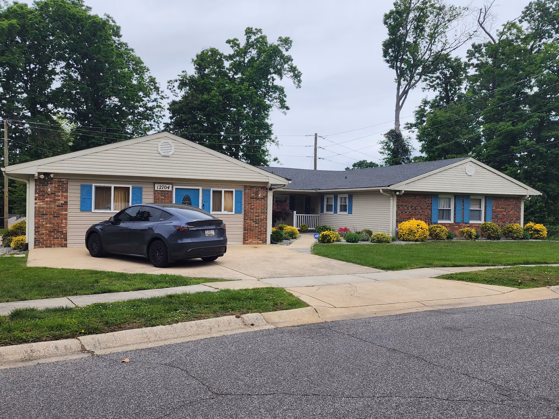Single-story ranch-style assisted living building with beige siding and brick, blue shutters, landscaped front yard and a dark car parked in the driveway.