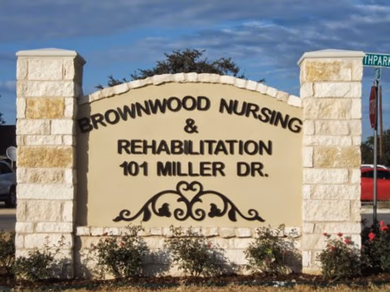 Stone sign for Brownwood Nursing & Rehabilitation located at 101 Miller Dr., with decorative black metal scrollwork at the bottom and some bushes planted in front. A street sign and parked cars are visible in the background under a partly cloudy sky.