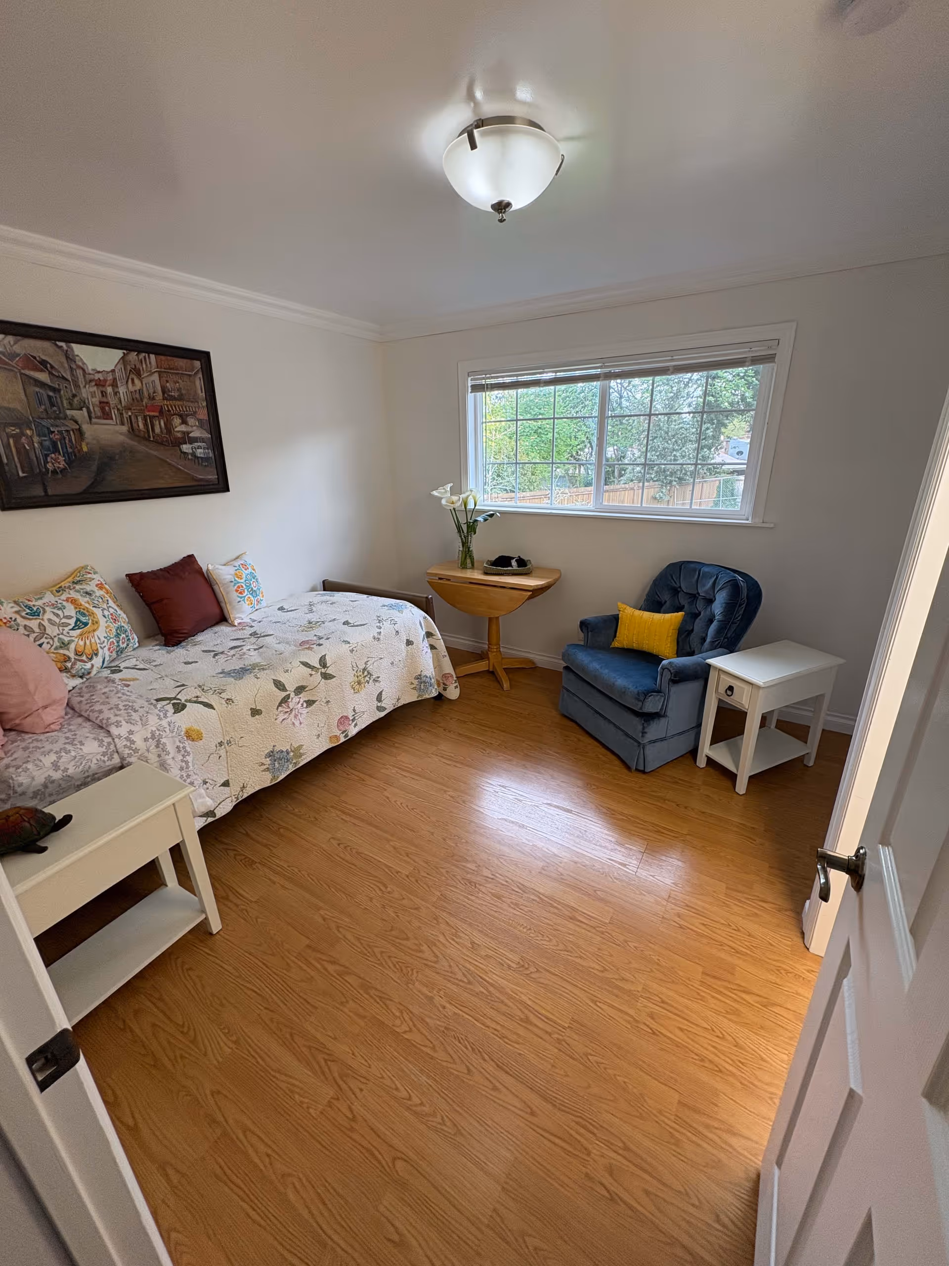 A cozy bedroom with a single bed covered in a floral quilt and multiple pillows. There is a small white nightstand next to the bed with a decorative turtle on it. A blue cushioned armchair with a yellow pillow is positioned near a window with blinds. A small wooden table with a vase of flowers and a pair of headphones sits between the chair and the window. The room has light-colored walls, wooden flooring, and a ceiling light fixture. A framed painting hangs on the wall above the bed.