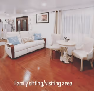 A cozy family sitting and visiting area with a white sofa adorned with decorative pillows, a small round table with a potted plant, and two upholstered chairs. The room has wooden flooring, light-colored curtains, and framed artwork on the walls.