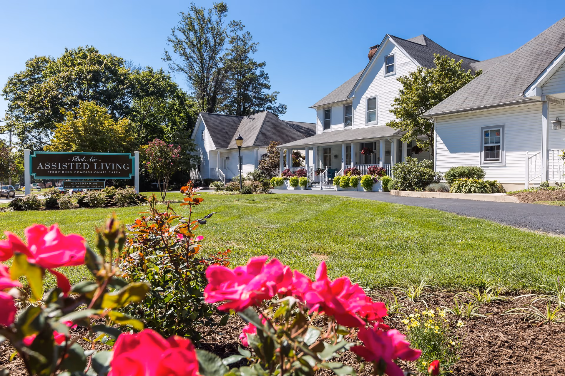 Exterior view of Bel Air Assisted Living facility with a well-maintained lawn, blooming pink flowers in the foreground, and white buildings with porches under a clear blue sky.
