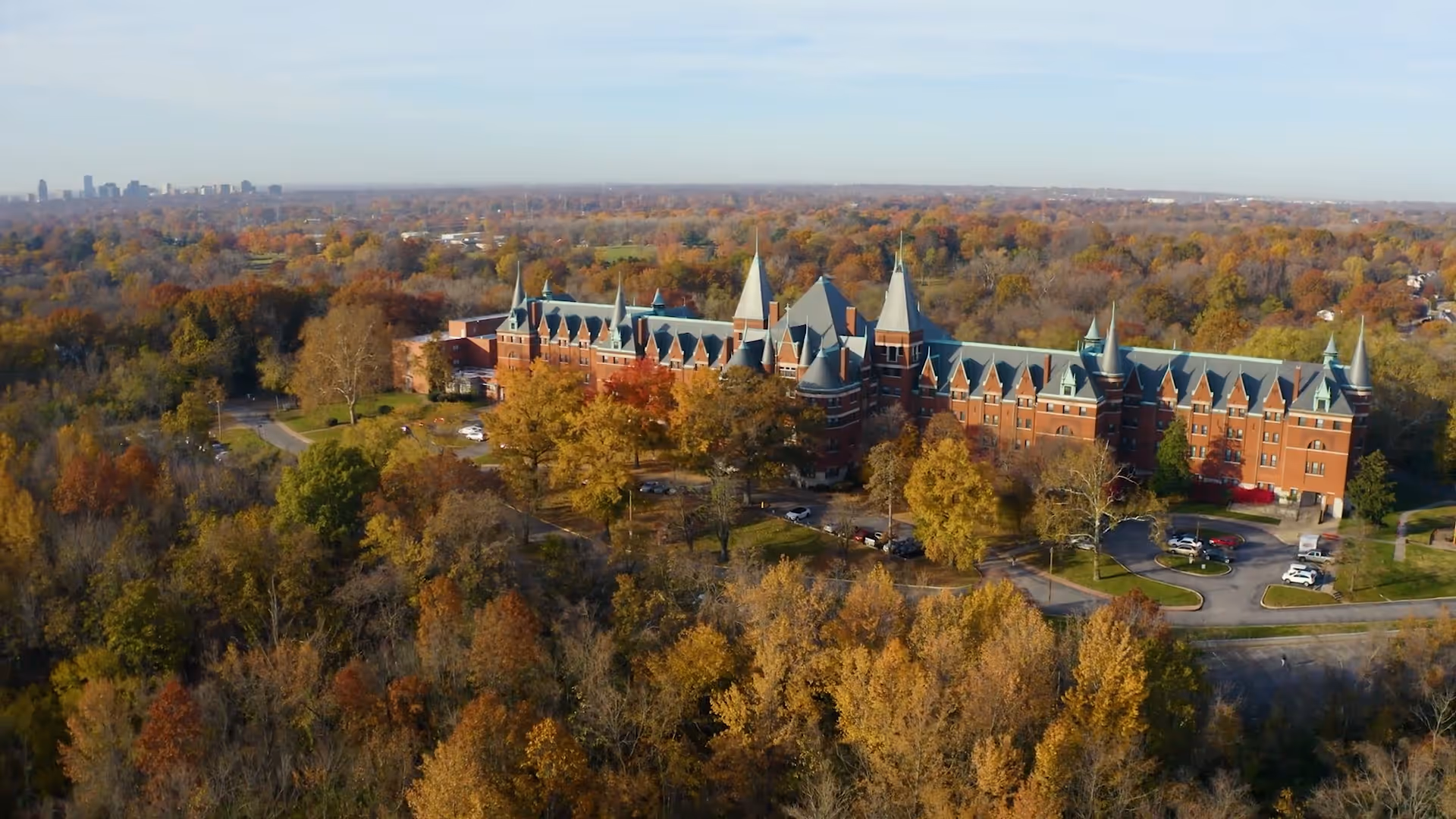 Aerial view of Normandy Nursing Center, a large brick building with multiple pointed towers and a green roof, surrounded by trees with autumn foliage and a parking area with several cars.