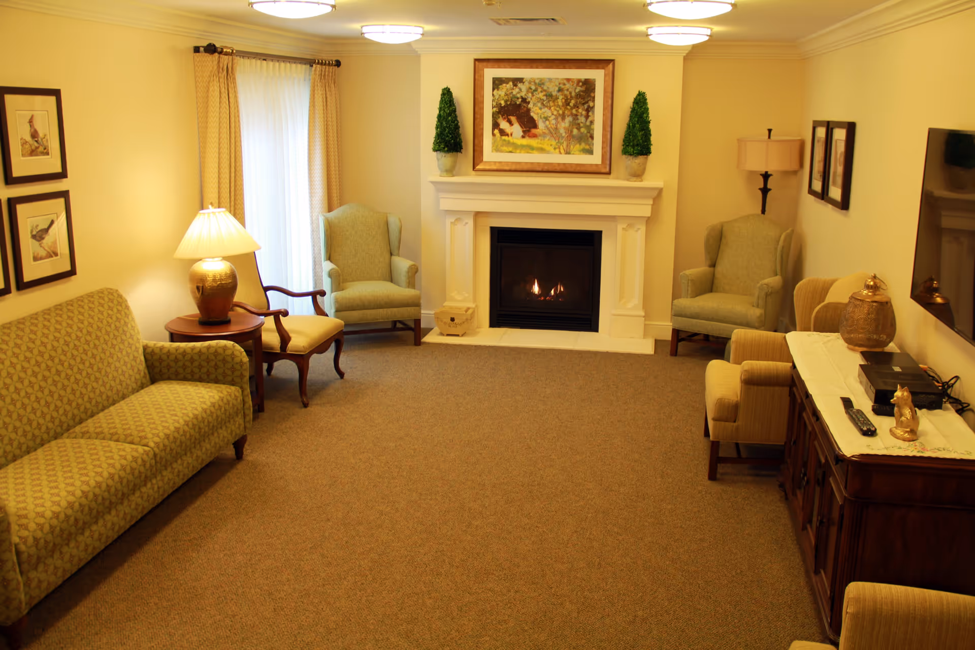 A cozy living room in a senior living facility featuring a lit fireplace with a painting and two small potted plants on the mantel. The room has several upholstered chairs and a sofa arranged around the fireplace. There is a wooden side table with a lamp, framed bird pictures on the wall, a wooden cabinet with decorative items, and a flat-screen TV mounted on the wall. The room is warmly lit with ceiling lights and has beige walls and carpeted flooring.