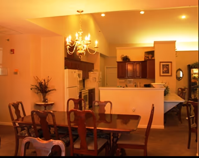 Dining area with a wooden table and chairs under a chandelier next to an open kitchen with refrigerator and cabinets.