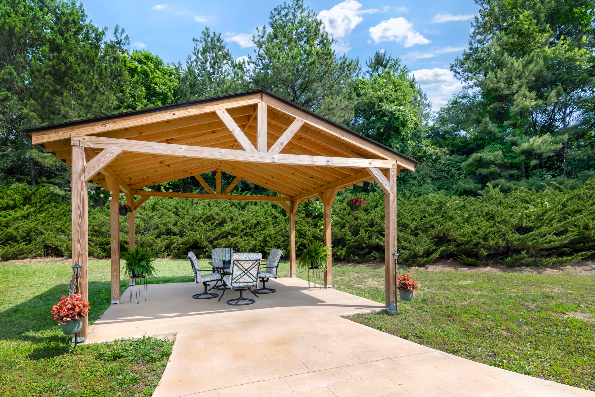 A wooden outdoor pavilion with a pitched roof situated on a concrete patio surrounded by green grass and trees. Under the pavilion, there is a glass-top table with six cushioned chairs. Hanging flower pots and potted plants decorate the pavilion posts.