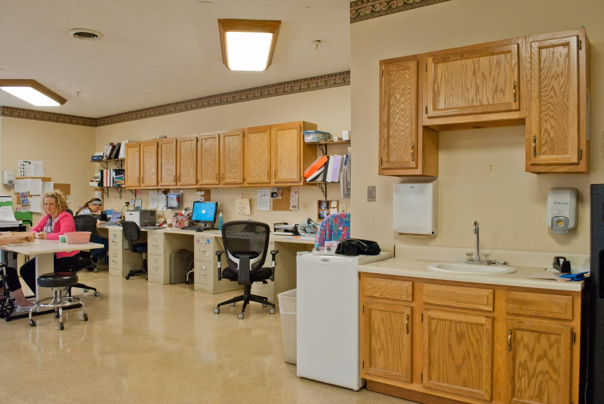 Interior view of an office area in a senior living facility with wooden cabinets mounted on the wall, desks with computers and office chairs, and a small kitchenette with a sink, mini fridge, and soap dispenser. Two women are seated at a table on the left side of the image.