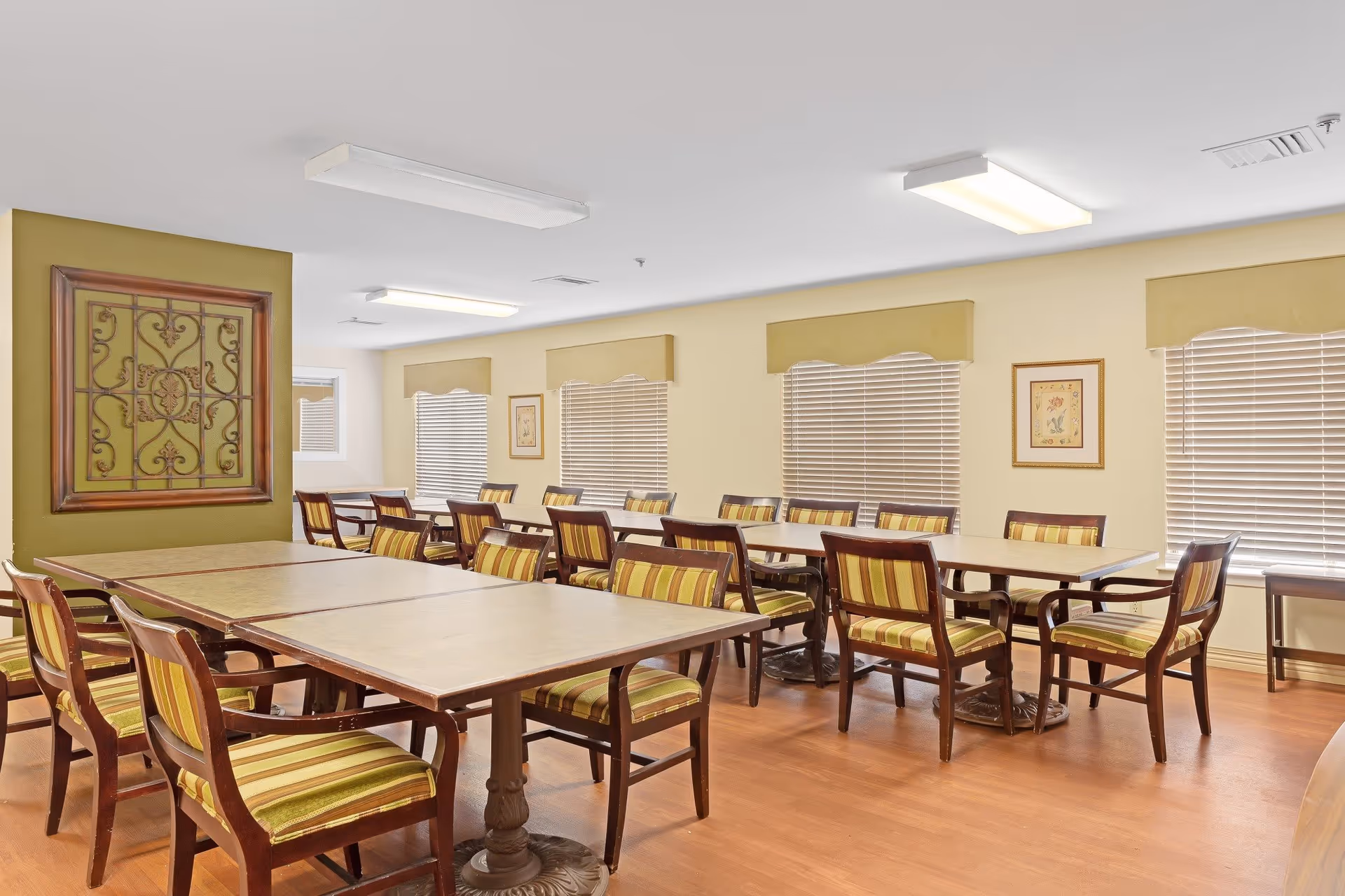 A bright dining room with multiple rectangular tables and wooden chairs with green and yellow striped cushions. The walls are light yellow with three windows covered by blinds and valances. There is a decorative framed metal wall art on one wall and a framed picture on another. The floor is wooden and the ceiling has fluorescent lights.