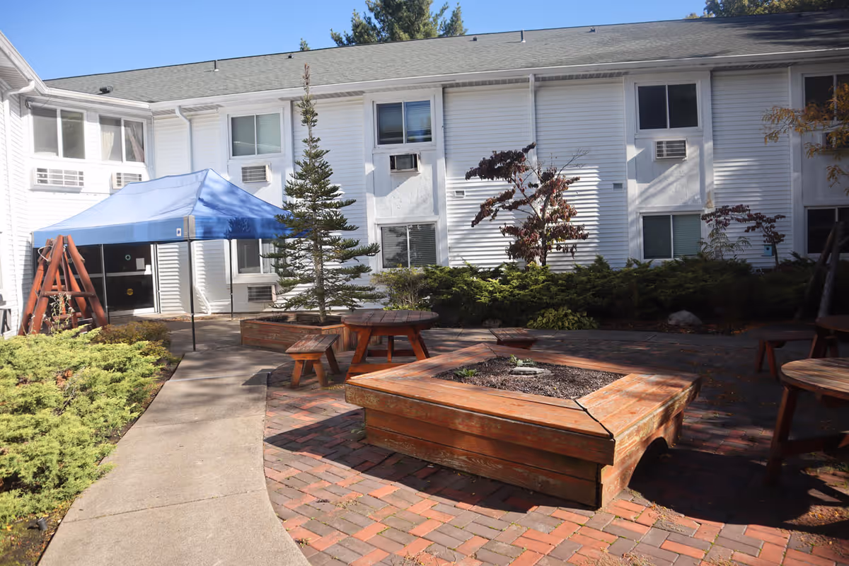 Outdoor courtyard area of a senior living facility with a paved walkway, wooden picnic tables, a wooden fire pit, a blue canopy tent, and various shrubs and small trees. The background shows a white two-story building with multiple windows and air conditioning units.