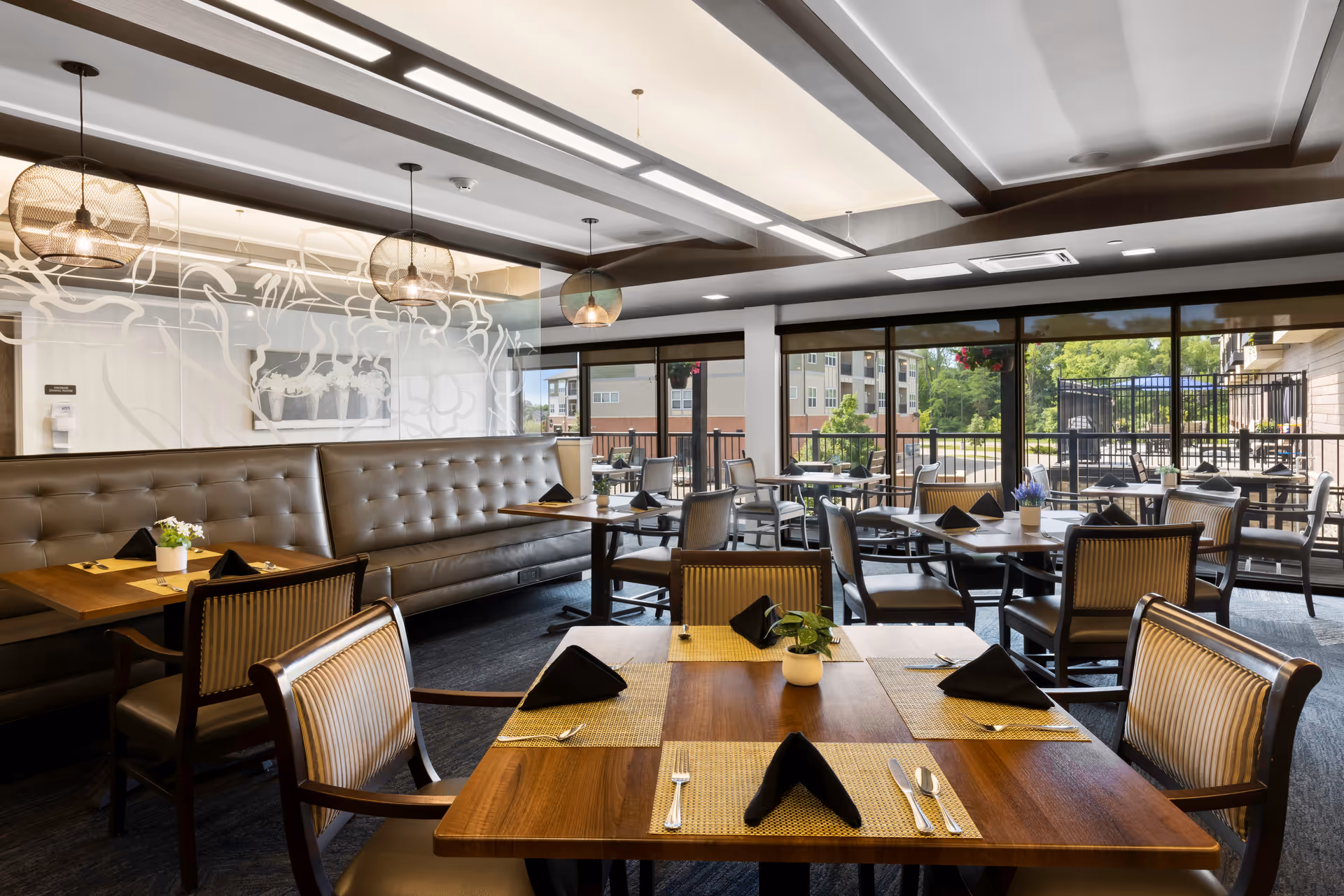 A bright and modern dining room with wooden tables set with black napkins, silverware, and small potted plants. The room features a long tufted leather bench along one wall, several chairs with striped upholstery, large windows letting in natural light, and pendant lights hanging from the ceiling. Outside the windows, there is a view of a patio area and neighboring buildings.
