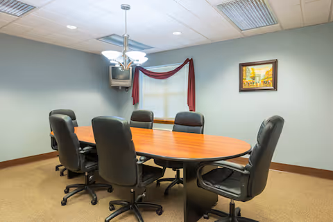 A small conference room with an oval wooden table surrounded by six black swivel chairs, a wall-mounted TV, window with a red valance, and a framed painting.
