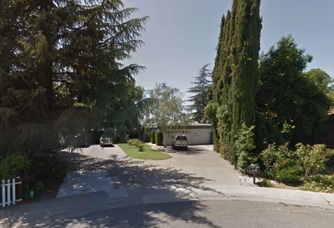 Exterior view of a facility with a driveway, parked cars, and tall trees.