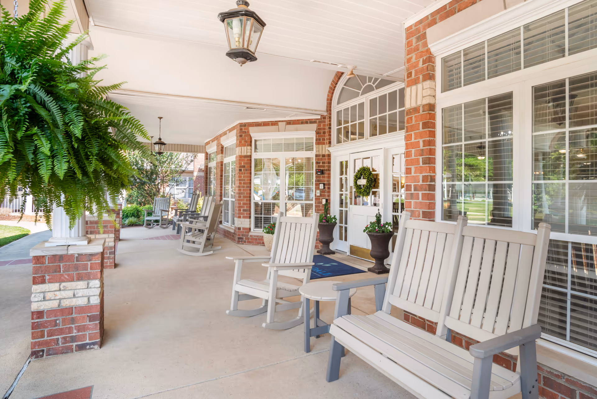 Covered porch area of a senior living facility with white rocking chairs and benches arranged along the brick exterior wall. Hanging green fern plants and potted flowers decorate the space. Large windows with white blinds and a white door with a green wreath are visible. The porch has a smooth concrete floor and ceiling-mounted lantern-style lights.