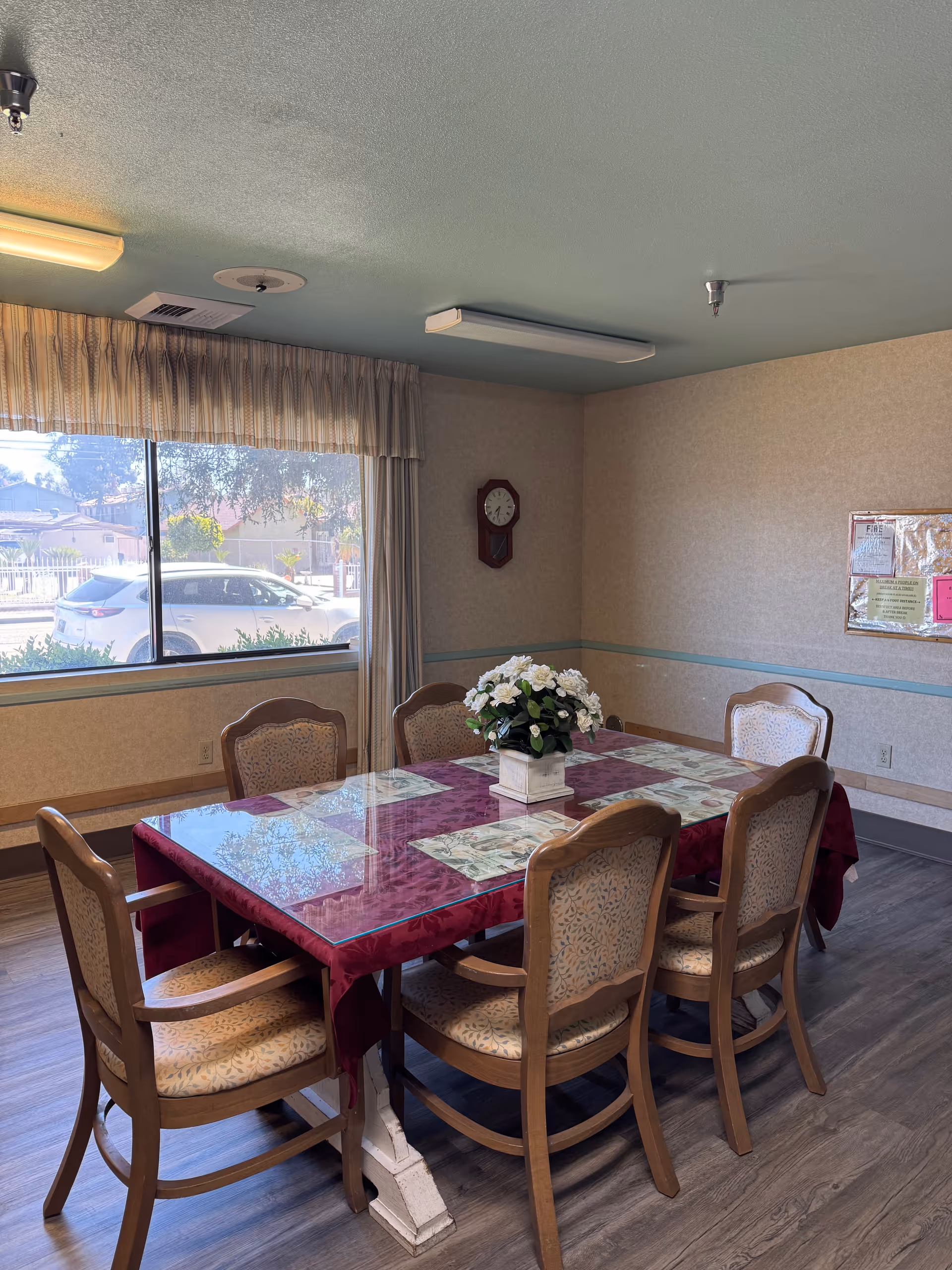 A small dining room with a rectangular table covered in a red tablecloth and placemats, surrounded by six wooden chairs and a floral centerpiece near a large window.