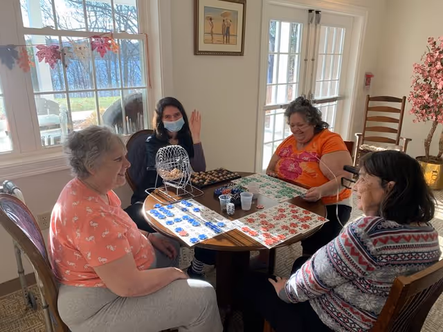 Four women sitting around a round wooden table playing a bingo game in a bright room with large windows and glass doors. One woman is wearing a face mask and waving, while the others are focused on their bingo cards. The room has light-colored walls, a framed picture, and a decorative pink flowering plant in the corner.
