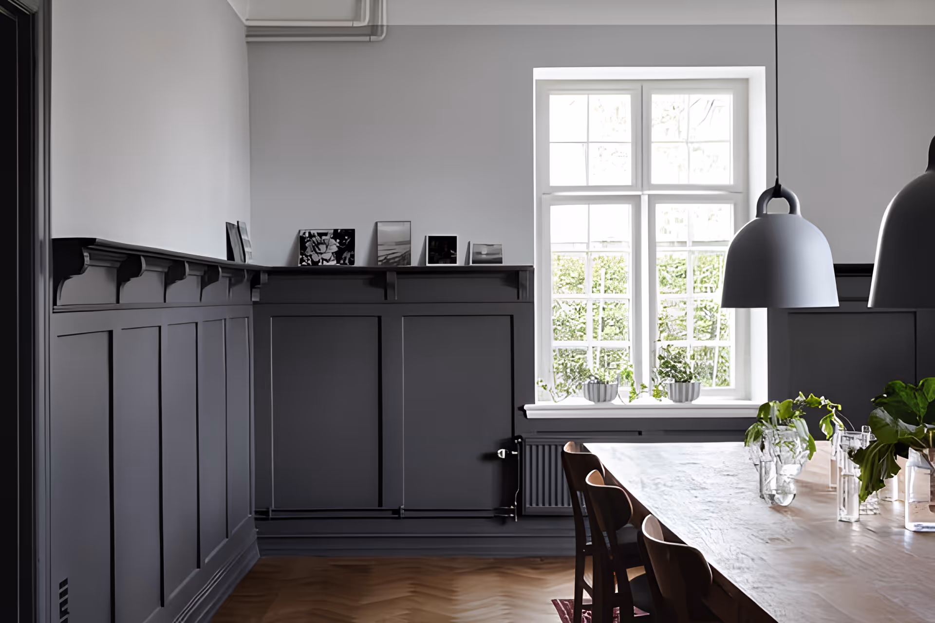 A bright dining room with a large wooden table surrounded by chairs. The room features dark gray wainscoting on the walls, a large window letting in natural light, and several small plants on the windowsill and table. Two modern pendant lights hang above the table.
