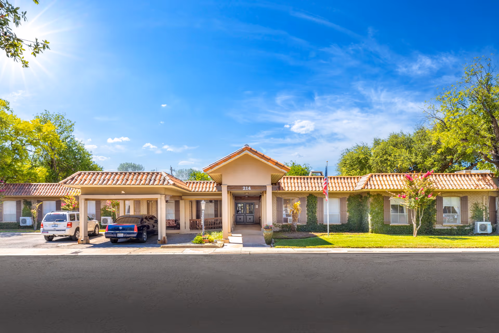 Front exterior view of 214 Martin-A Retirement Residence building with a tiled roof, two cars parked in front, an American flag on the lawn, and trees surrounding the property under a clear blue sky with the sun shining.