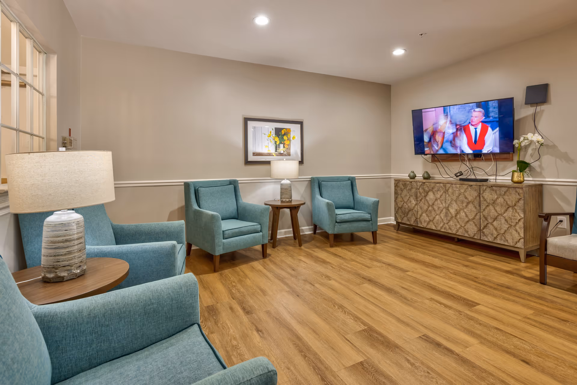 A cozy living room with four teal armchairs arranged around two wooden side tables with lamps. A flat-screen TV is mounted on the wall above a decorative wooden cabinet. The room has wood flooring, beige walls, and a framed picture hanging on the wall.