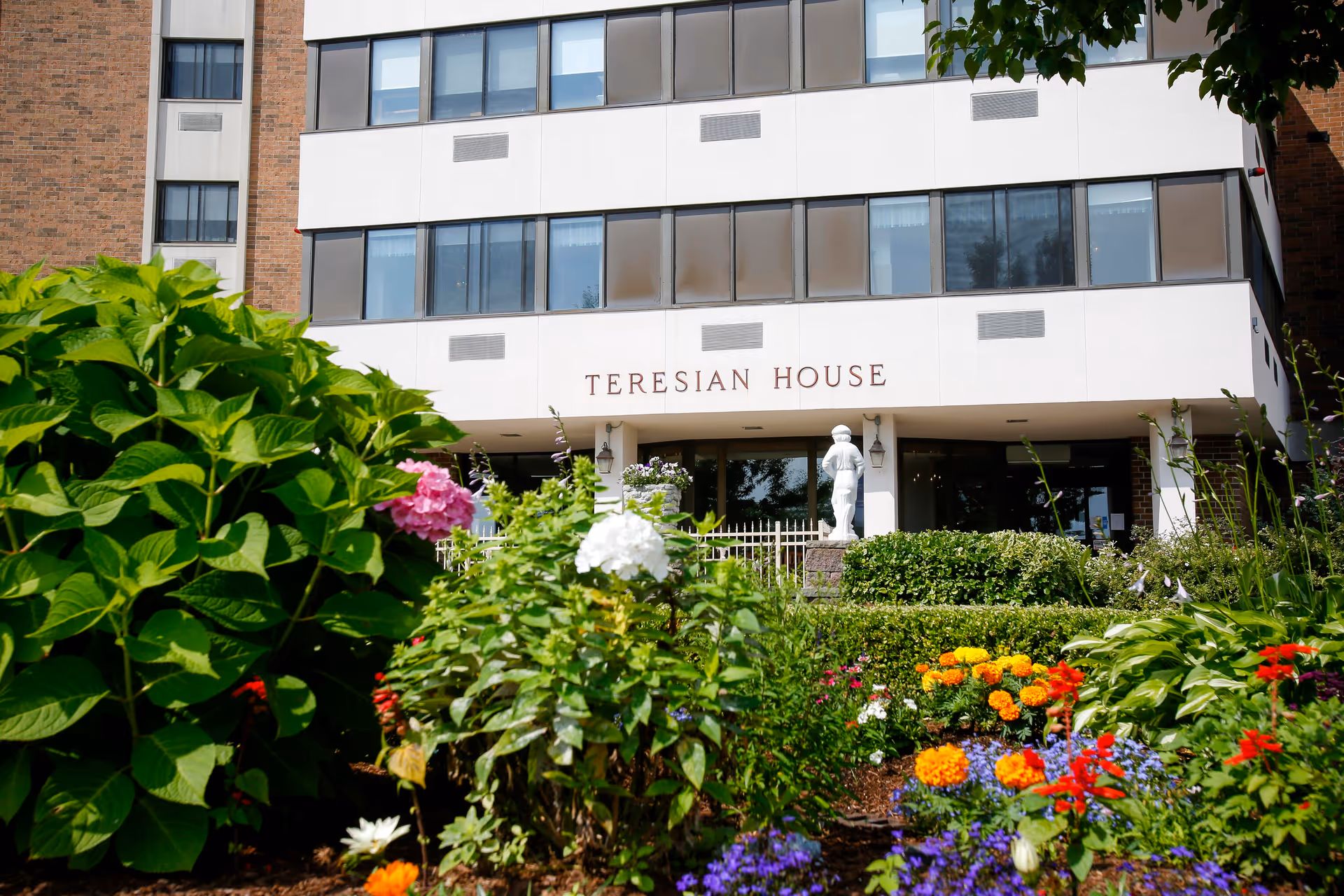 Front exterior view of Teresian House Center for the Elderly building with multiple windows and a garden with colorful flowers and green bushes in the foreground.