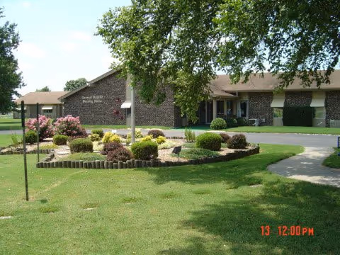 Exterior view of General Baptist Nursing Home building with a brick facade, surrounded by a well-maintained lawn, landscaped garden with bushes and flowers, and a large tree providing shade.
