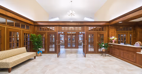 Spacious wood-paneled reception lobby with a tiled floor, glass double doors, seating bench, potted plants, and a front desk under a chandelier.