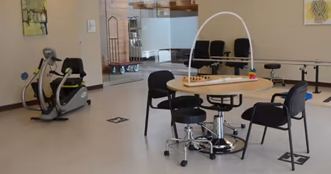 A therapy or rehabilitation room with exercise equipment including a stationary bike, a round table with chairs around it, and a handrail along the wall. The room has light-colored walls and flooring, with a glass door and some chairs visible in the background.
